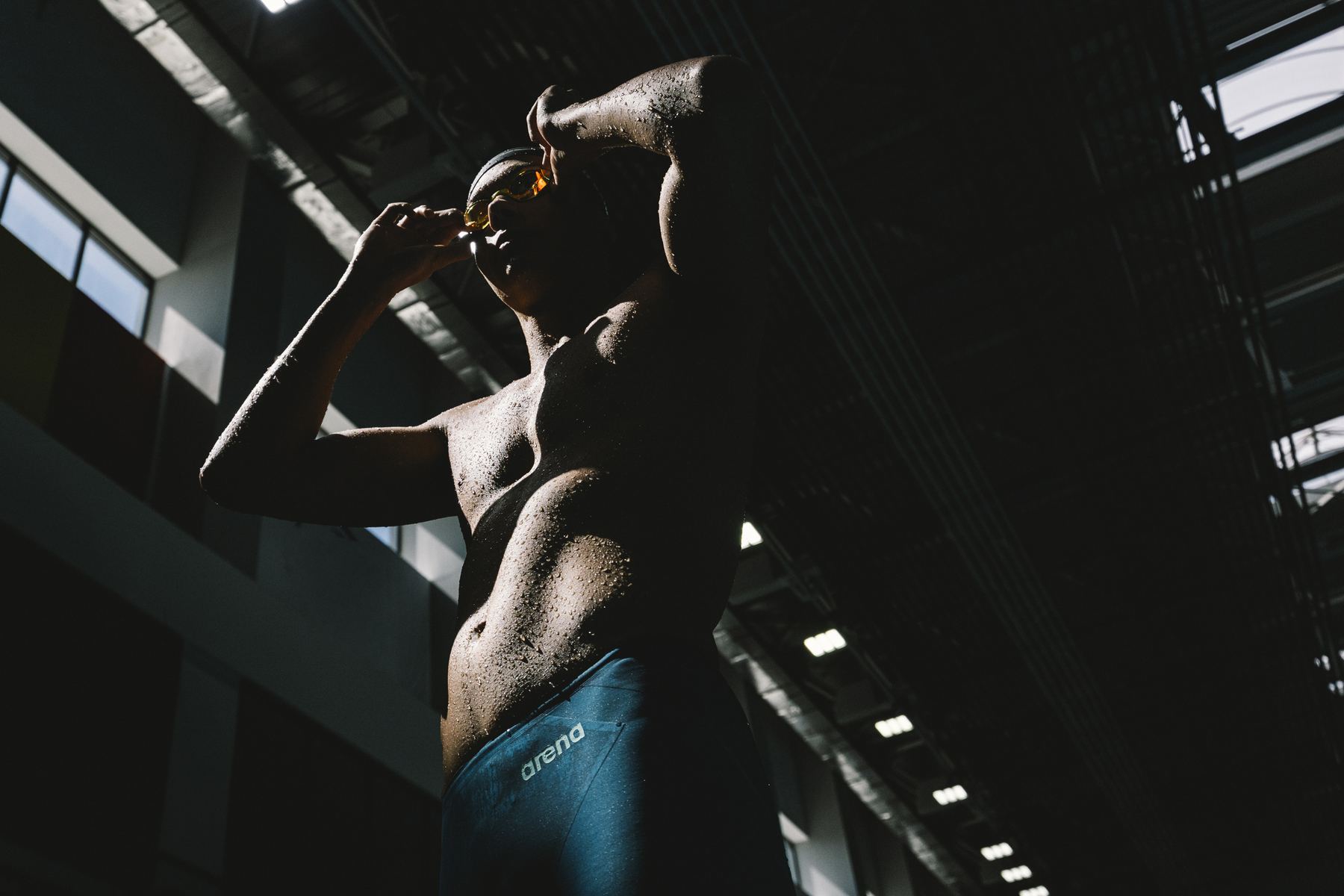 Swimmer in the Arena Powerskin AIR3 jammer fixing goggles in a dark pool hall, lit by a single shaft of light